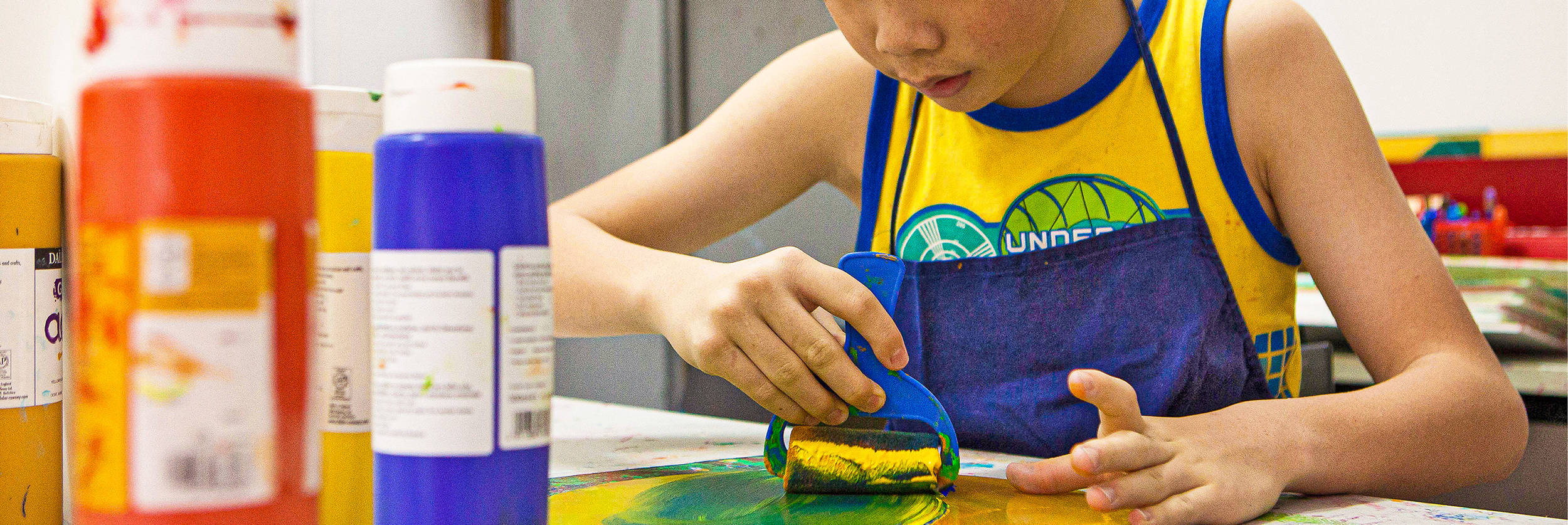 his banner features a young student using a roller brush to apply green and blue paint onto a canvas.