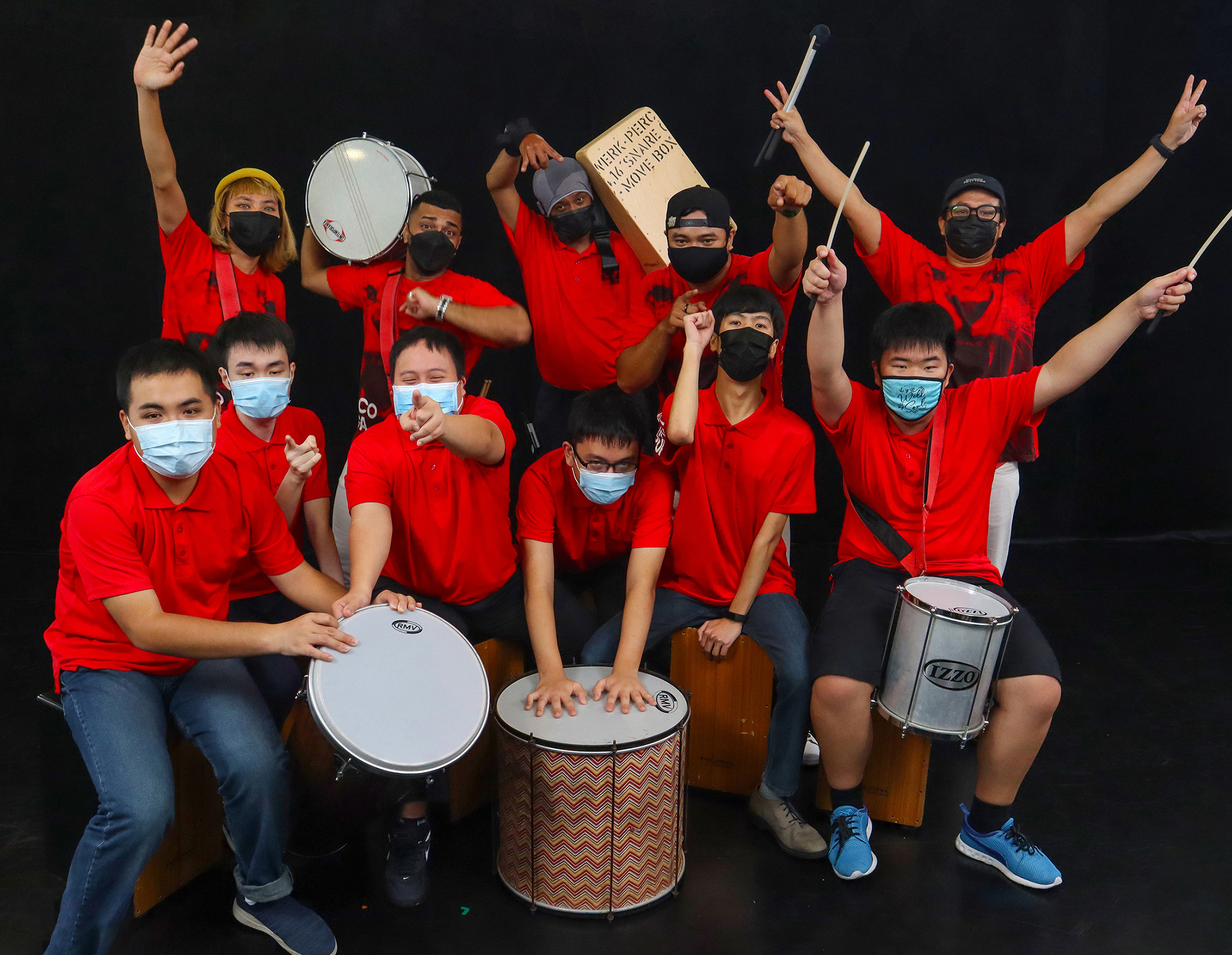This image features six of ART:DIS’s cajon students posing with their teacher and four musicians from Bloco Singapura. The students are each sitting on their instruments and cheering at the camera, while the adult musicians are all raising their drums/batons and fist-pumping into the air. 